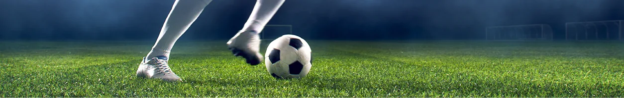 Fotografía panorámica de las piernas de un jugador conduciendo un balón sobre césped, con el arco desenfocado al fondo y sin rostros a la vista.