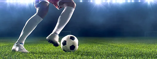 Recorte de la misma escena de fútbol nocturno, con el balón detenido sobre el pasto junto a la zapatilla del jugador.